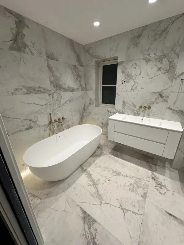 Large-format marble-effect wall and floor tiles around a freestanding bath with brass taps
